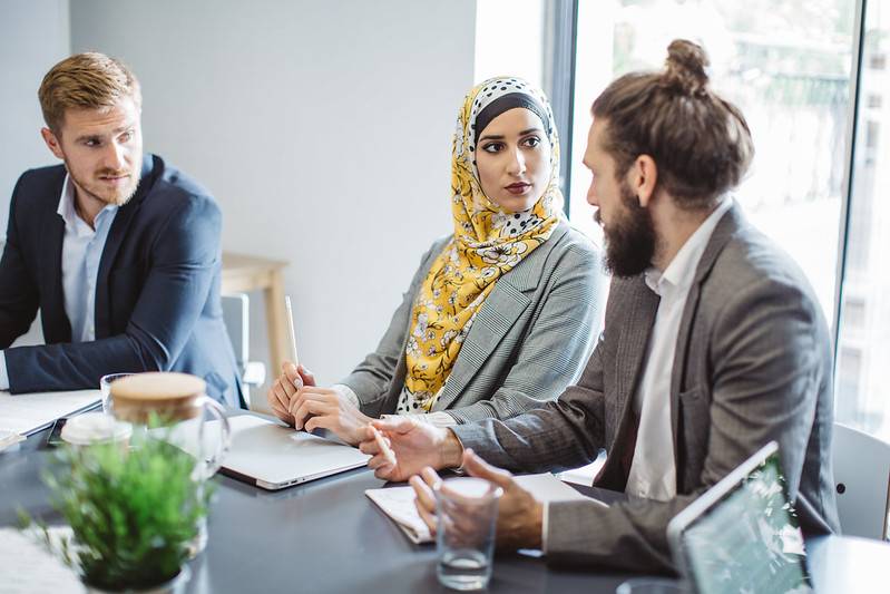 To menn og ei kvinne med hijab snakker saman rundt eit møtebord